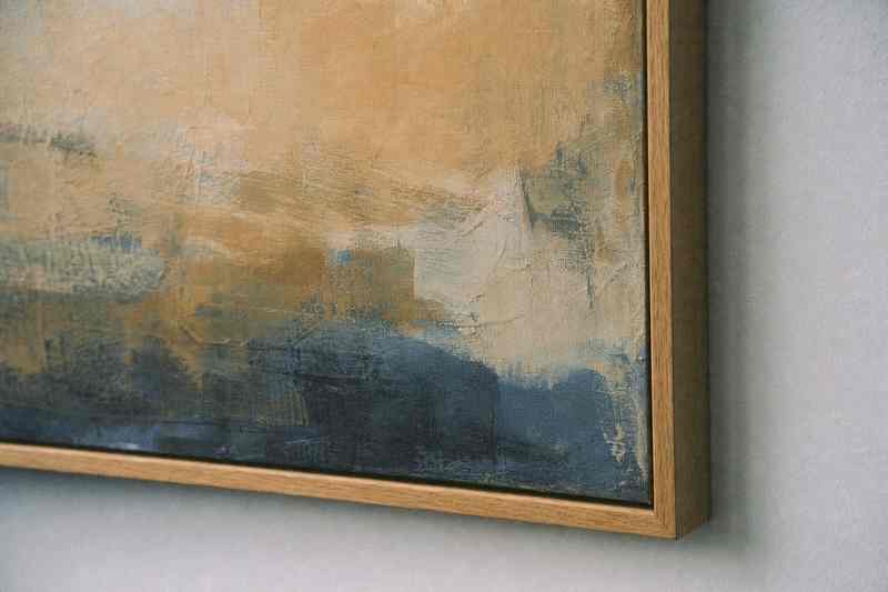 De hoek van een ingelijst abstract schilderij in oker en blauw aan een lichte wand